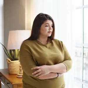 A Woman Holds Her Arms as She Looks Out the Window of Her Home
