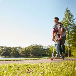 A dad rides a scooter with his son on a sunny day in the park.