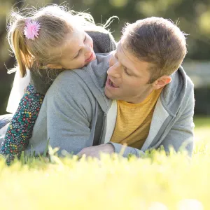 A little girl climbing on her fathers back laying down in the grass.