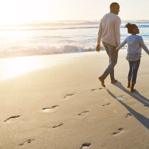 A family of three walks along the beach at sunset leaving footprints in the sand.
