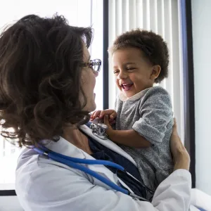 A female pediatrician holds a smiling infant in an exam room.