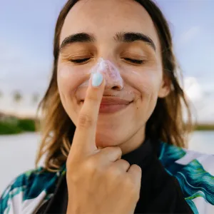 A woman on a beach wearing sunscreen on her nose and touching her nose with her index finger.