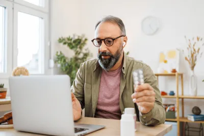 Man attending a virtual care appointment at home on laptop holding medication.