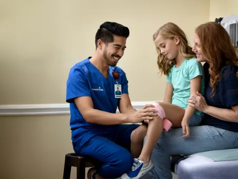 A Provider Wraps a Child Patient's Knee With Pink Gauze While a Parent Watches