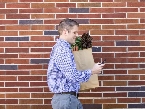 A young Caucasian man checks his cell phone while carrying a bag of fresh produce.