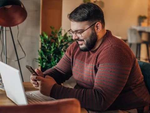 A Man Smiles as He Uses a Cellphone While Sitting at a Desk in front of a Laptop.