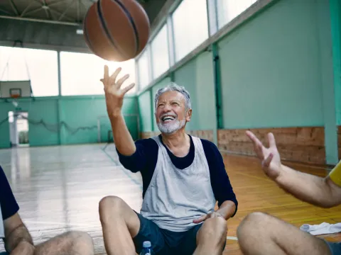 A Trio of Senior Men Sit on a Gymnasium Floor While One of Them Tosses a Basketball in the Air