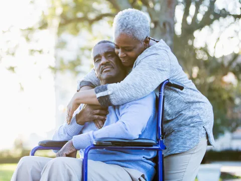 A Woman Embraces Her Husband as He Sits in a Wheelchair Outdoors in a Park