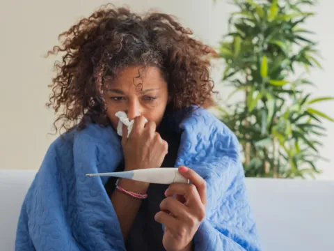 A Woman Blows Her Nose While Sitting on Her Couch at Home Reading a Thermometer 