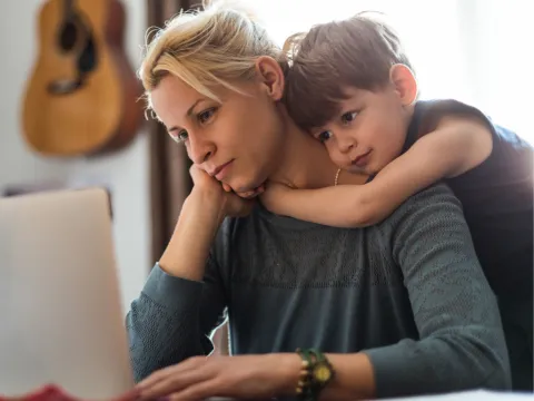 A Mom Stares at A Computer Screen While Her Young Son Hugs Her From Behind