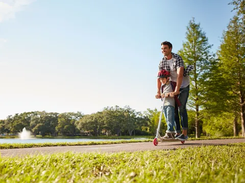 A dad rides a scooter with his son on a sunny day in the park. 