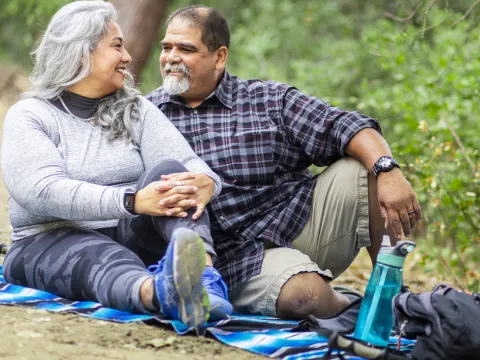 A senior couple is sitting on the ground outdoors enjoying a picnic after a hike.