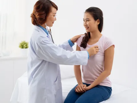 A woman is sitting on an exam table while a doctor listens to her heart.