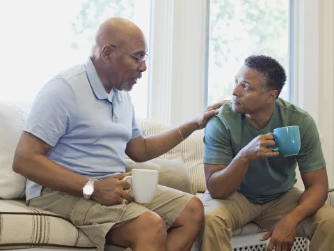 An African American father and son chat with coffee mugs on the living room couch.
