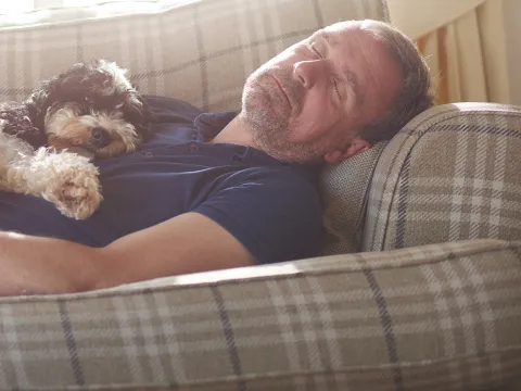 A middle aged man asleep on a patterned sofa with a white and black curly haired dog on asleep on top of him.