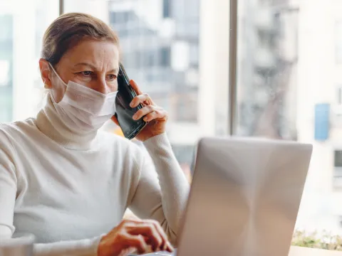 Mask wearing woman talking on the phone while using a laptop indoors.