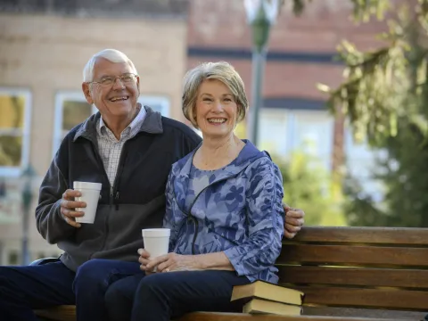 Elderly couple sitting on a park bench drinking coffee.