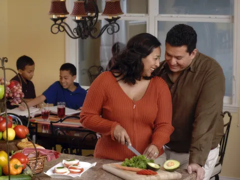 A couple shares a sweet moment while the wife prepares dinner, as their kids do homework in the background