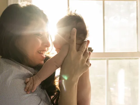 A woman stands in front of a window, holding up a newborn baby and smiling as she looks in his eyes.