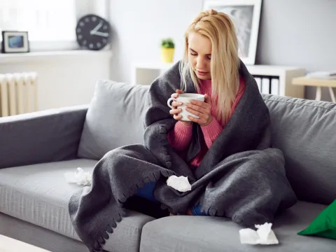 A woman drinking tea under a blanket at home.