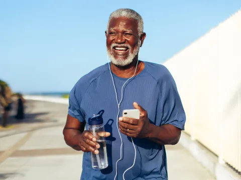 An older black man using headphones plugged into his phone while walking outdoors and holding a water bottle.