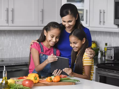 A mother and her 2 young daughters reading a recipe on a tablet in the kitchen.