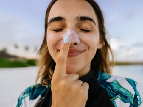 A woman on a beach wearing sunscreen on her nose and touching her nose with her index finger.