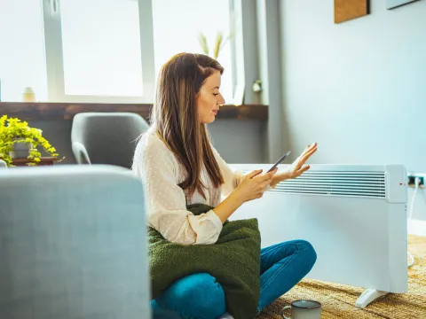 Woman using her phone while sitting next to a space heater at home.