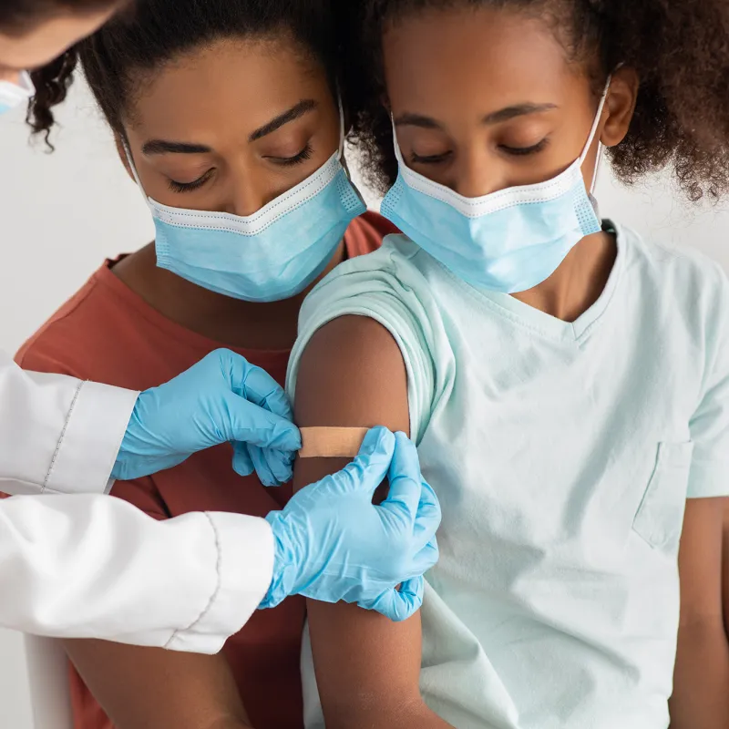A Mother Comforts Her Daughter While a Physician Puts a Bandaid on an Injection Site in the Upper Arm.
