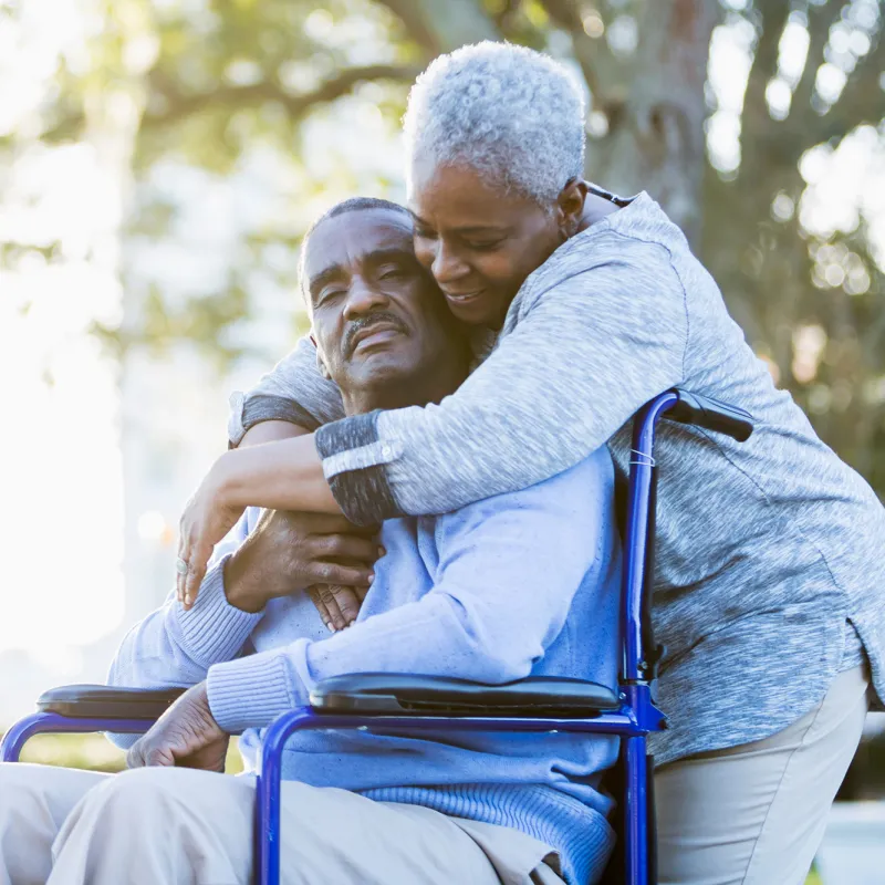 A Woman Embraces Her Husband as He Sits in a Wheelchair Outdoors in a Park