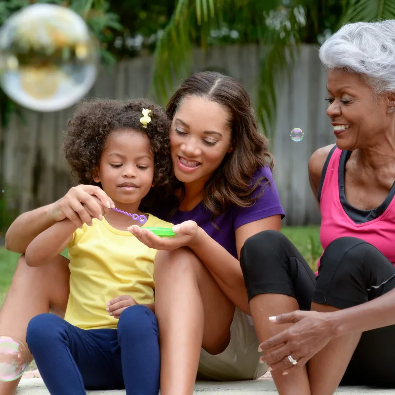 A grandmother, mother, and daughter play with bubbles outside. 
