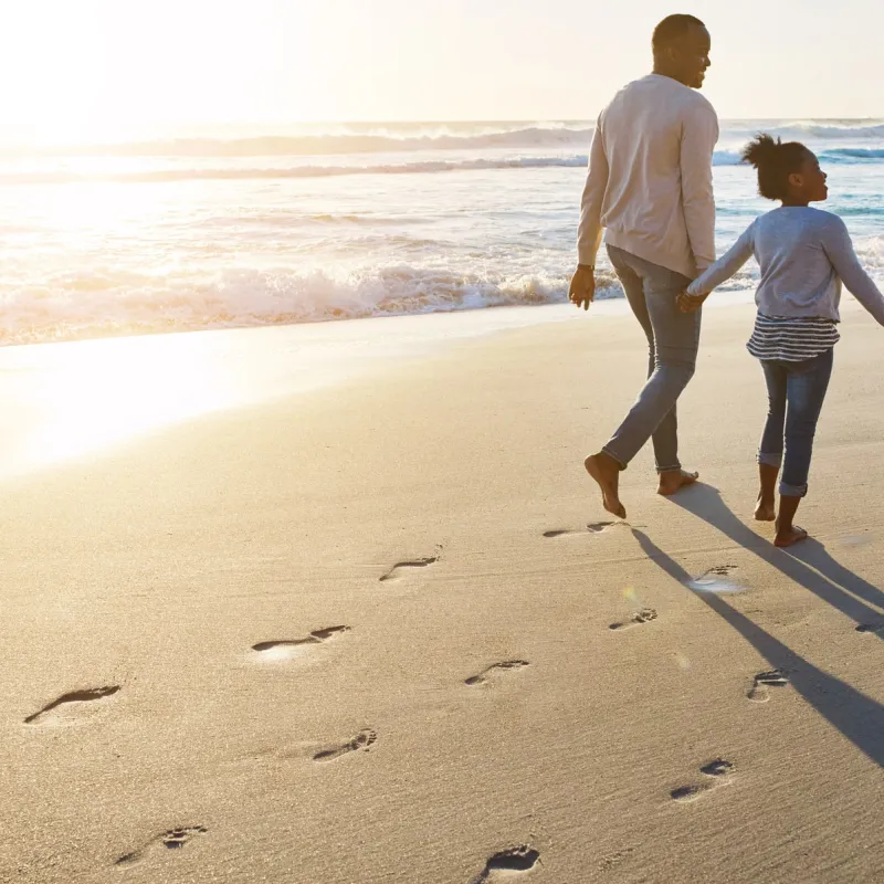 A family of three walks along the beach at sunset leaving footprints in the sand.