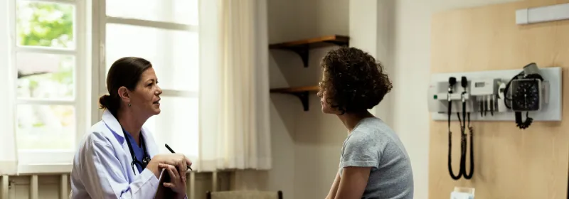 A young woman sits in a hospital room and talks about her health and wellness with her primary care doctor.