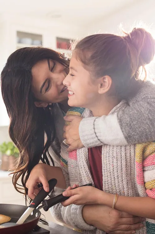 A family of three women are in the kitchen, one daughter hugging the youngest while she flips pancakes.