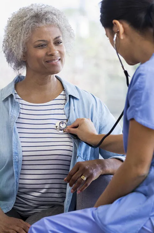 A female doctor is seated, listening to a senior woman's heart with a stethoscope.